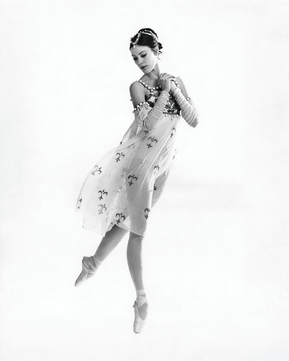 A young Kain in a flowy transparent dress with a pattern and details at the top. She has her hair up in a headpiece. She clutches her hands together, is up on pointe on one foot and has the other leg behind her in a tiny arabesque. She looks down towards her foot