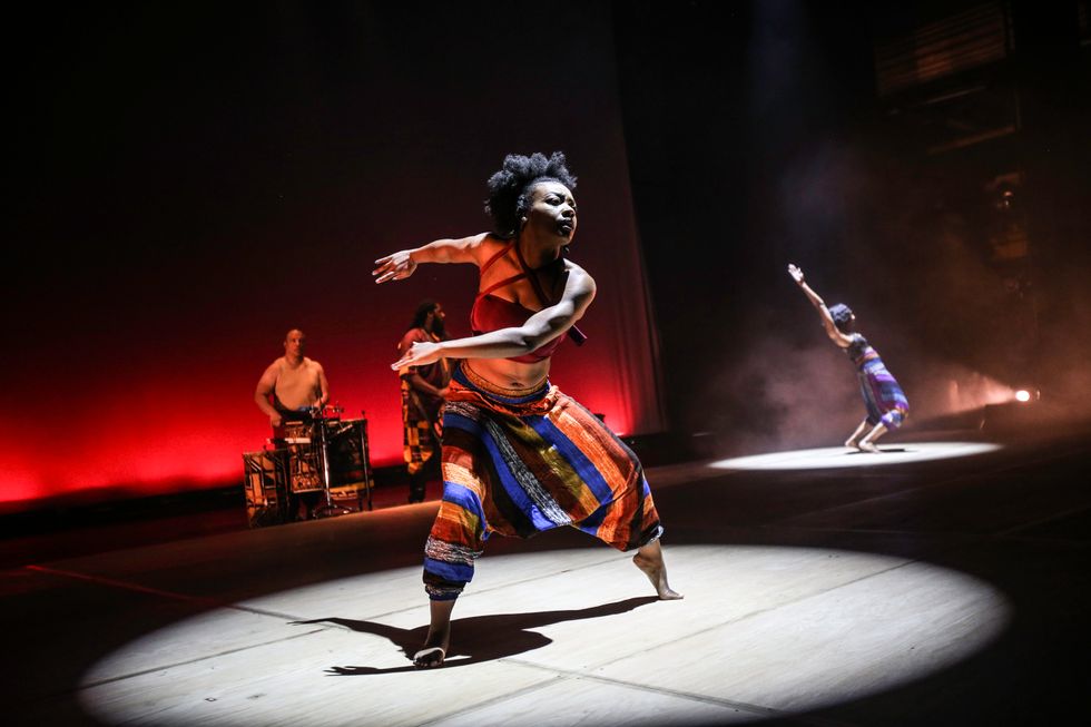 In a spotlight, a toned black woman in brightly colored, traditional pants and a red crop top moves through a deep pliu00e9, her left arm crossing in front of her torso and right arm swinging back as though winding up. A musician is visible upstage, and another woman hinges in a spotlight in the background.