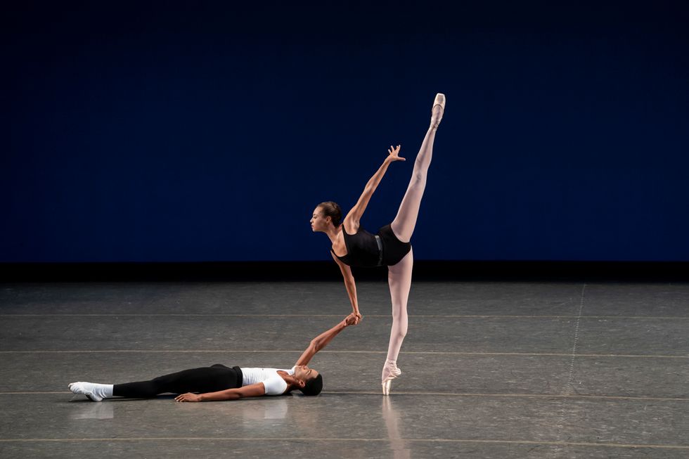 A male dance student in black tights and a white t-shirt lays down on the stage, holding the right hand of a female dance student as she balances in penchu00e9 on pointe.