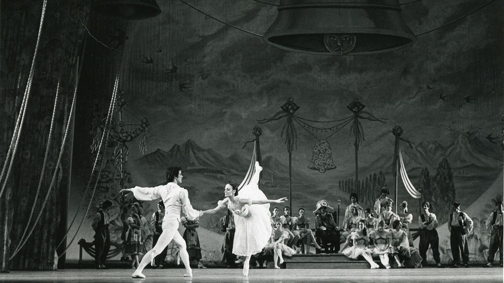 In this black and white photo, a man in a white ballet costume lunges and holds the hand of a ballerina in a long tutu as she balances in penchu00e9 croisu00e9.