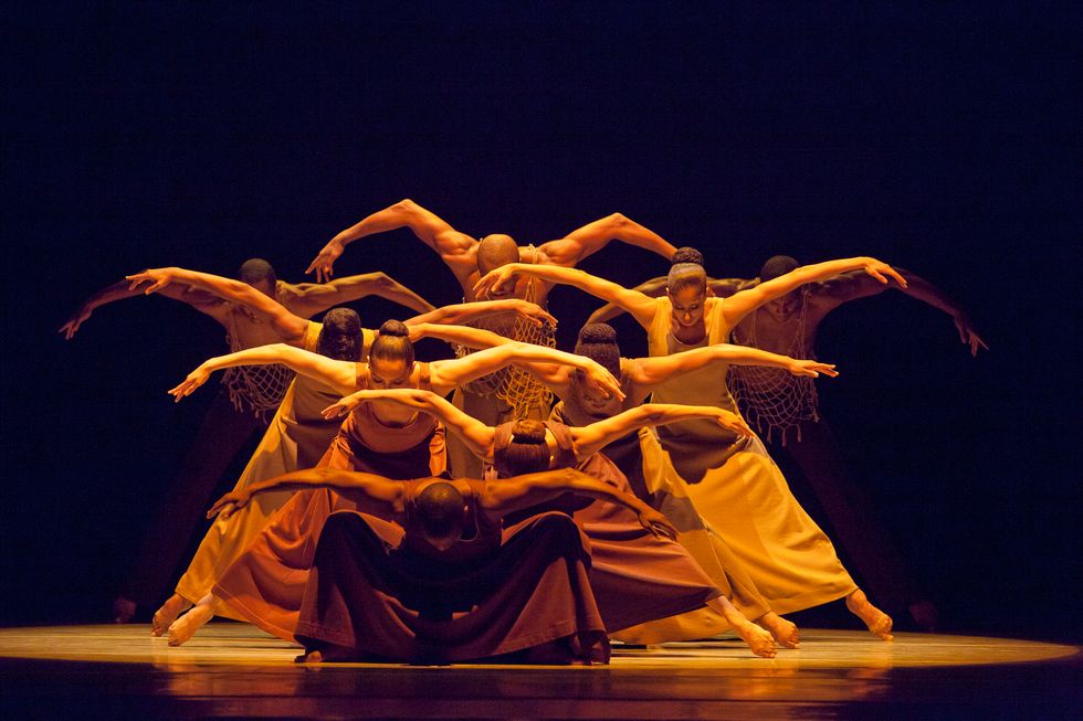 A group of dancers in a bright spotlight lunge down with their heads down and arms spread in second position.