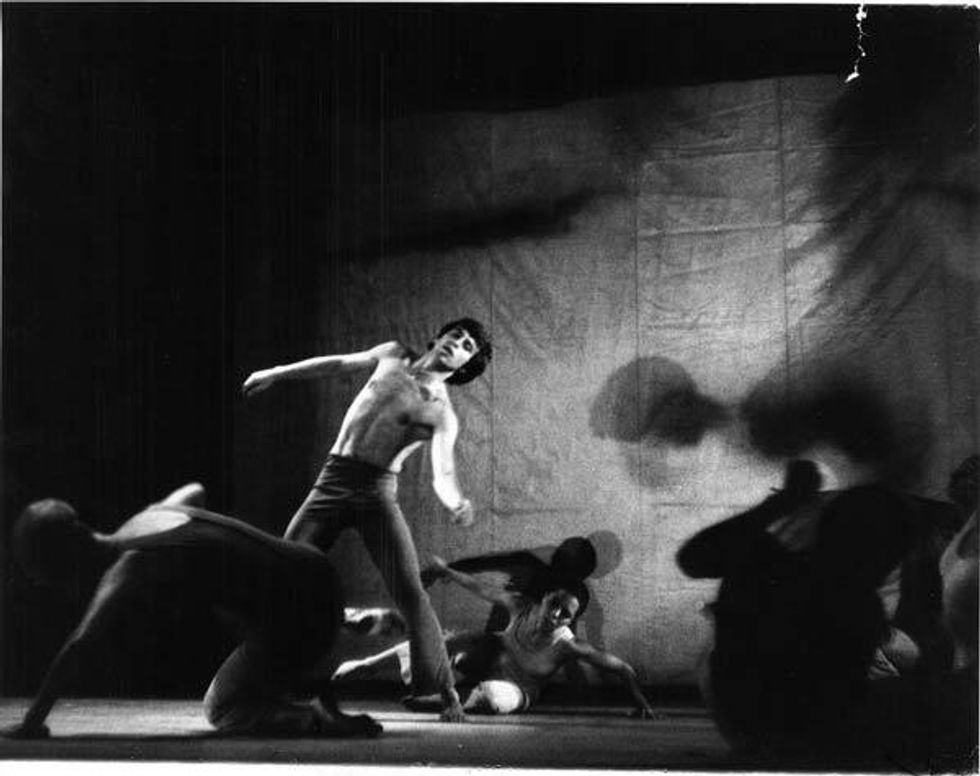 Black and white image of dancers in front of a wall with shadows. Some stand, some crawl on the floor.