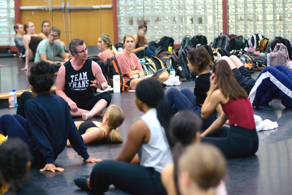 Dorsey sits at the front of a classroom, wearing a black "team trans" tank top. Students sit and lay on the floor around him, listening as he talks.
