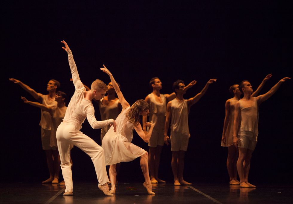 In the foreground a male and female dancer, both wearing white, have their backs to the camera. They stand in pliu00e9, with their right foot to the side in forced arch. They lean to their right, their right elbows bending gently at the elbow as their left stretches on diagonal upwards to complete the line. Blurry int he background, seven similarly dressed dancers stand with their feet together, facing the downstage diagonals, outside arms raised to eye level.