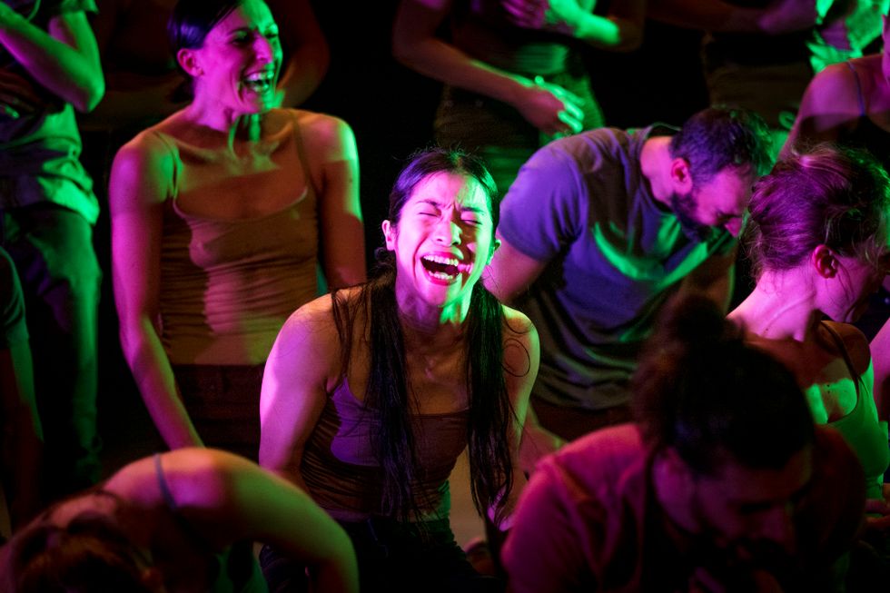 A woman with long, dark hair contorts her face in either a laugh or a grimace. She is in a crowd of out-of-focus, but similarly affected dancers. All are bathed in a mixture of purple and green light.