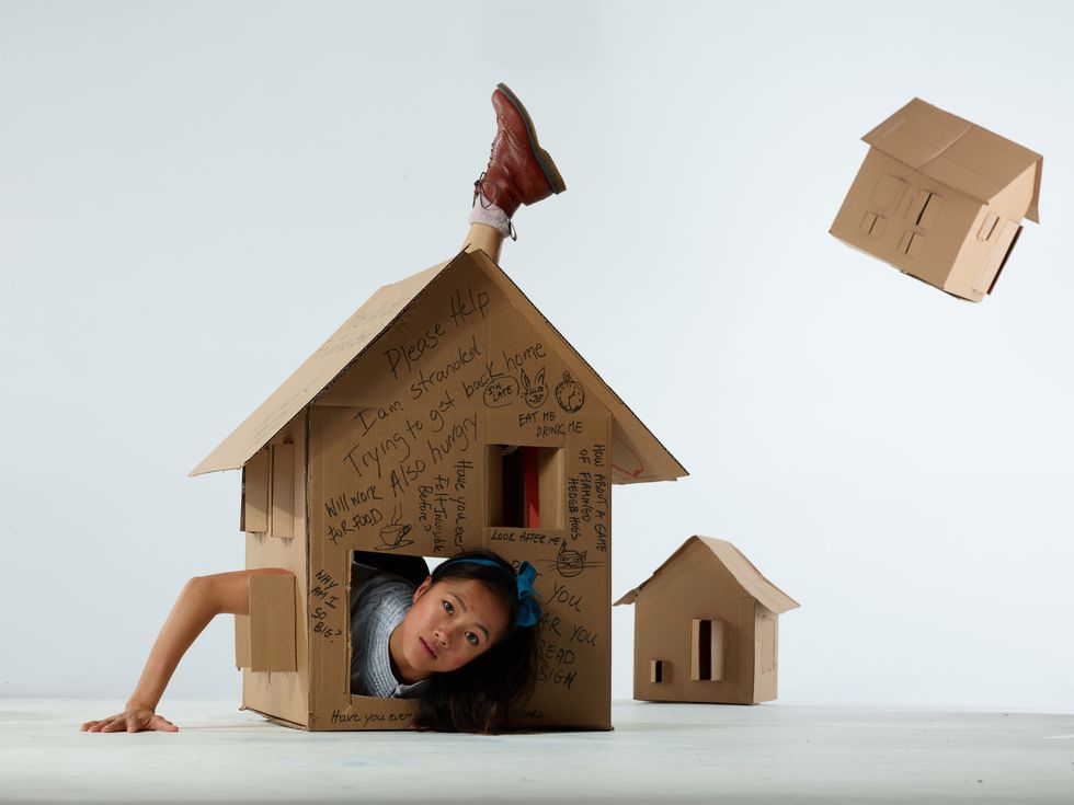 A woman with a blue ribbon in her dark hair pokes her head out of an opening in a cardboard box made to be shaped like a house, her right arm peeking out of another window to press against the floor, a booted foot stretching out of the back to show over the roof. The box has messages scrawled in black that read, "Please help. I am stranded. Trying to get back home. Also hungry." Another, smaller cardboard house is seen in the background, and a third flies through the air to the right.