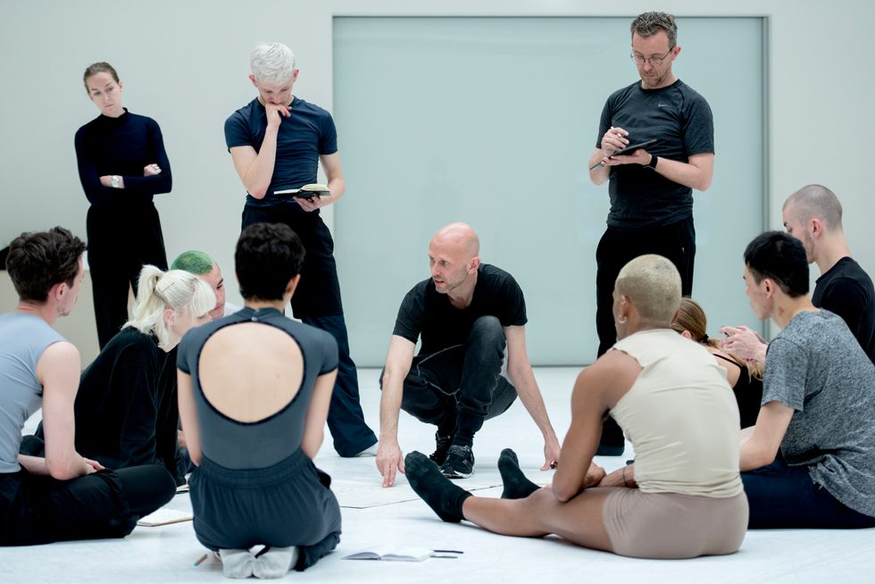 Dancers in rehearsal clothes sit in a circle, listening to a man crouching at the center of a white studio, pointing with two fingers at a point on the floor as he gives direction. Three others stand slightly behind him, looking intently at the notebooks or tablets held in their hands.