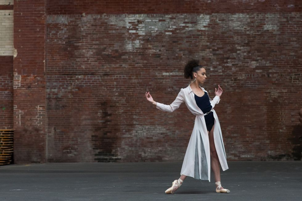 Chyrstyn Mariah Fentroy stands in front of a brick wall wearing a black leotard and long overcoat. Looking towards her left, she opens her arms and bends her elbows slightly while beveling over her right pointe shoe.