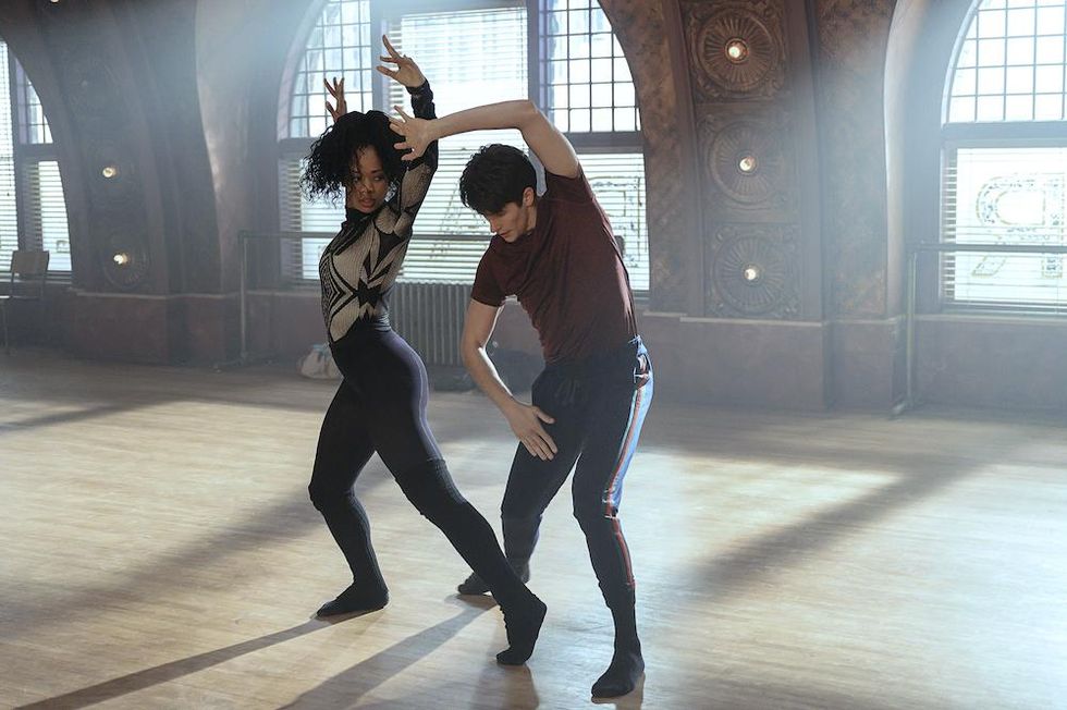 A still from "Tiny Pretty Things." Kylie Jefferson and Barton Cowperthwaite dance together in an empty, sun-lit studio. Kylie Jefferson's arms are extended above her head, and her legs are bent, with one extended to the side, on the floor. She wears black leggings and a metallic, geometric-printed long sleeve shirt. Barton Cowperthwaite stands with both legs bent, and one arm extended over his head. He wears navy joggers and a maroon tee shirt.