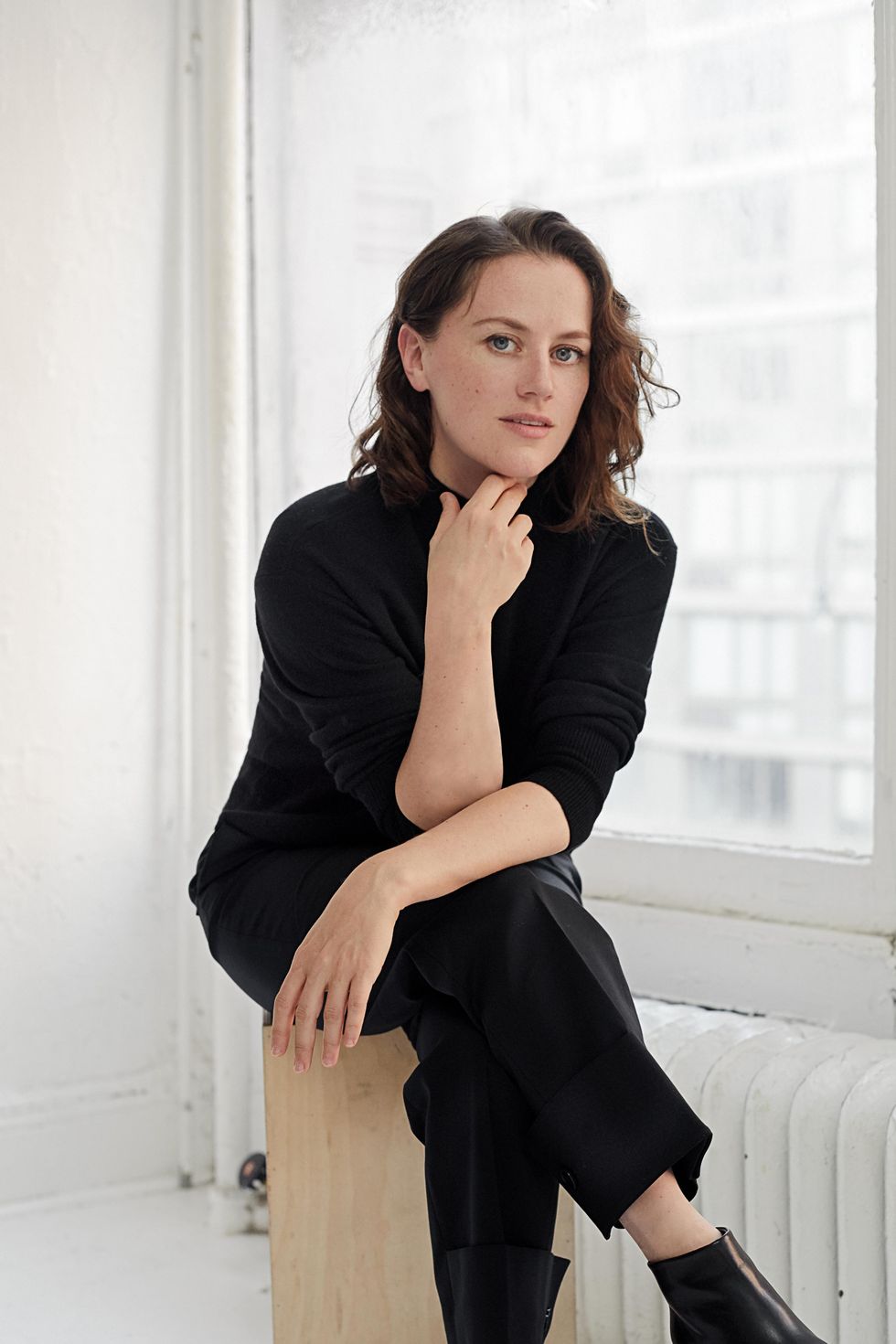 Helen Simoneau, a young, white brunette woman, looks questioningly at the camera, dressed in all black as she sits with her legs crossed, a hand caressing her chin.