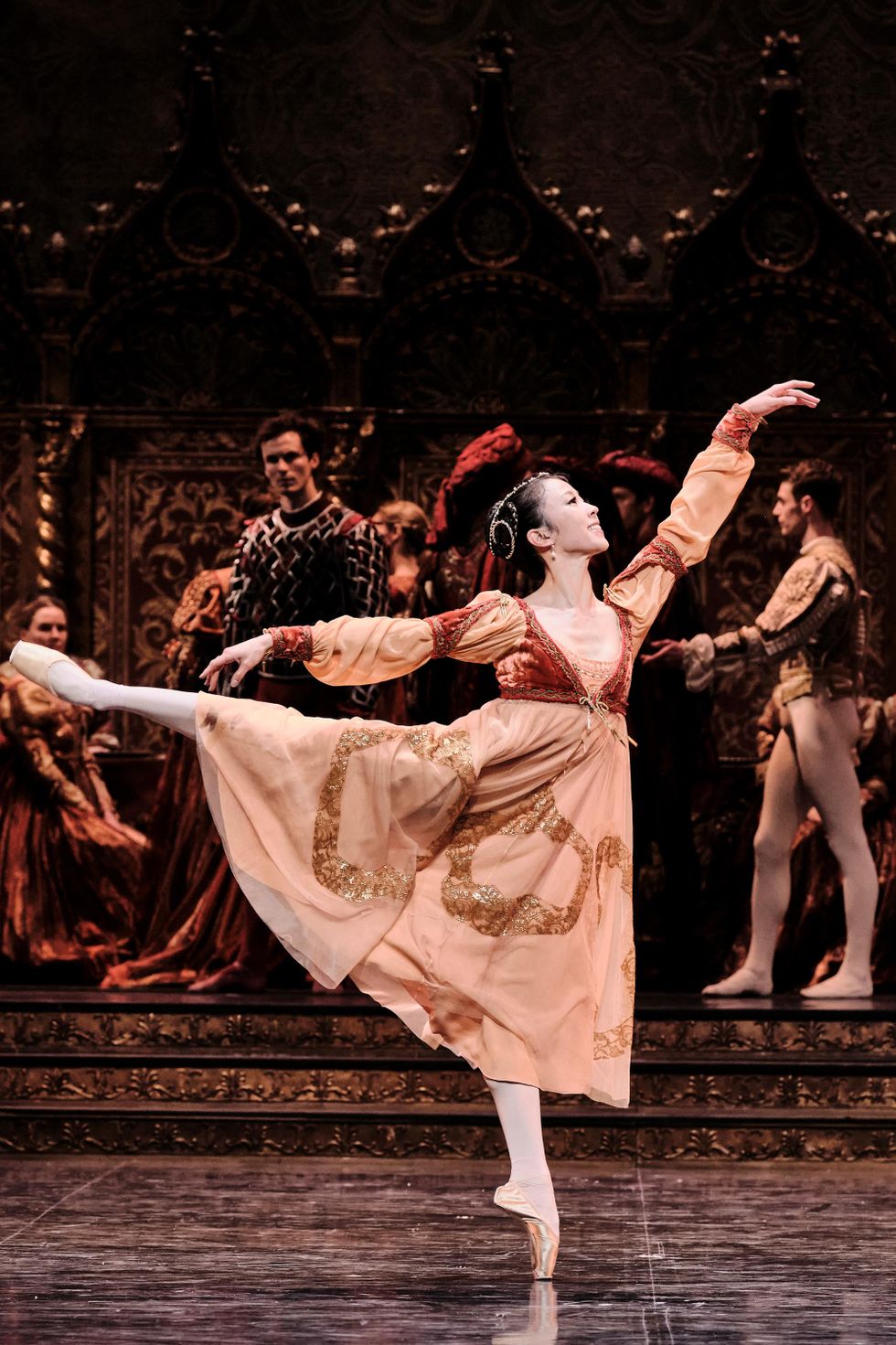 Sae Eun Park, in an elaborate peach and rust dress, pink tights, and pointe shoes, smiles as she balances in first arabesque. Behind her, dancers in opulent dresses and tunics watch.