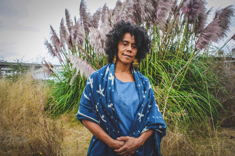 Amara Tabor-Smith stands with her hands clasped in front of her, a calm, questioning look directed towards the camera as a swath of plant life sways in the wind behind her.