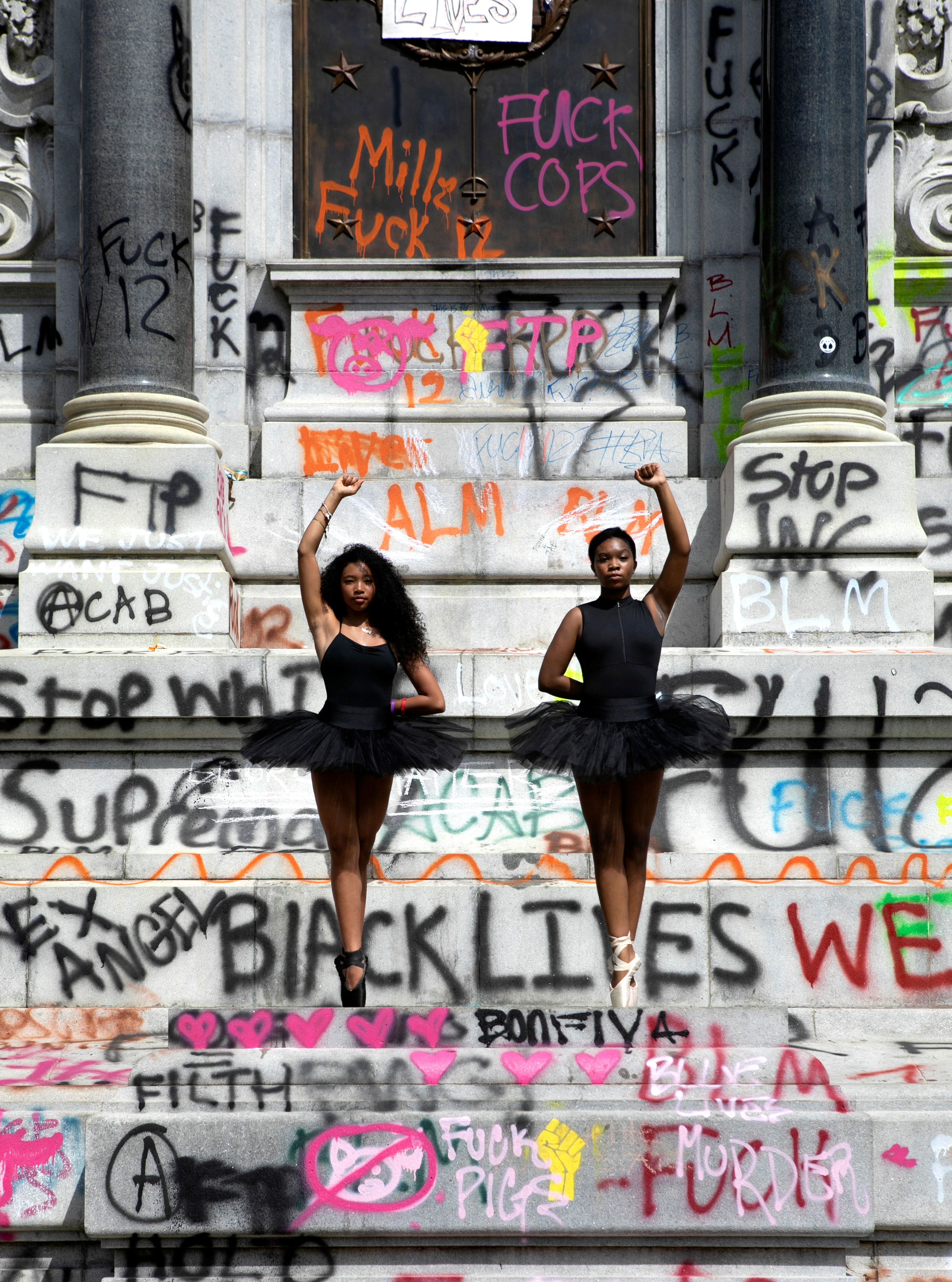 Two teenaged Black ballerinas pose in fifth position en pointe, each dancer raising a fist. They are standing on a graffiti-covered Confederate monument. The dancers are wearing black leotards and black tutus.