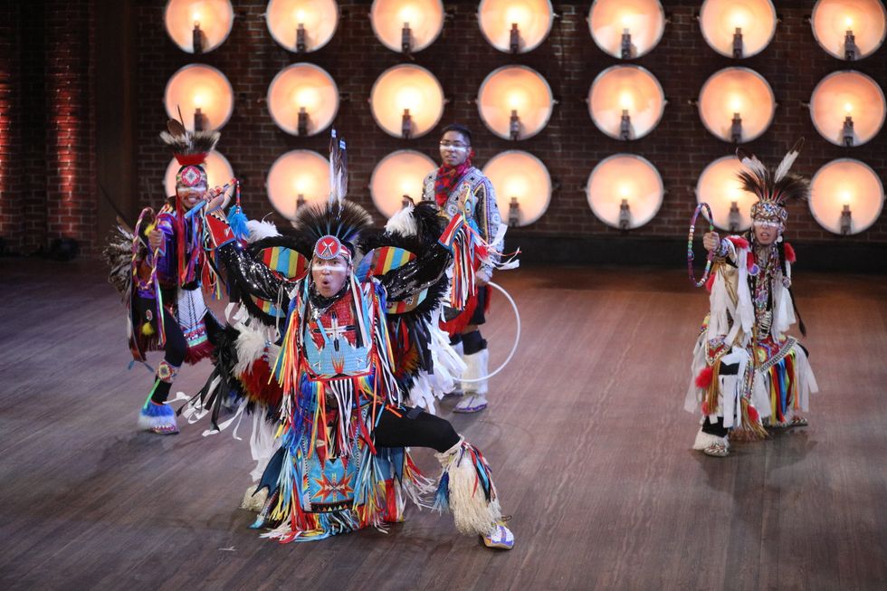 Four men dancing Native American powwow dances in colorful traditional costumes. Three hold traditional hoops.