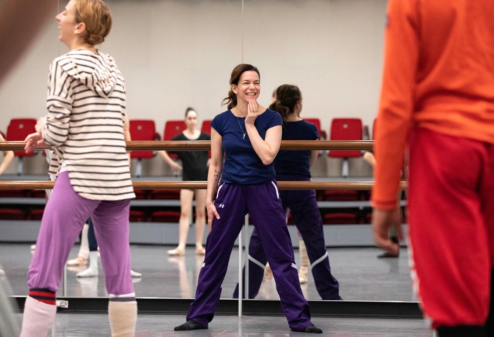 Pam Tanowitz stands in a second position in front of a barre and mirrors, smiling at dancers walking in front of her.