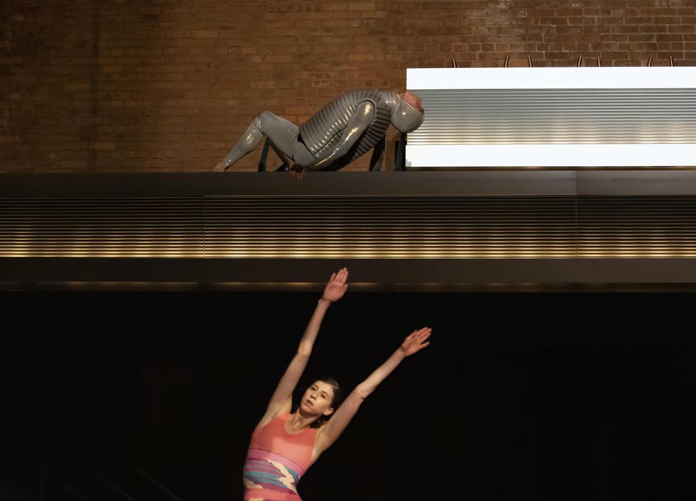 A dancer in silver leans back on all fours on stage, while a woman in pink below him reaches her arms diagonally side.