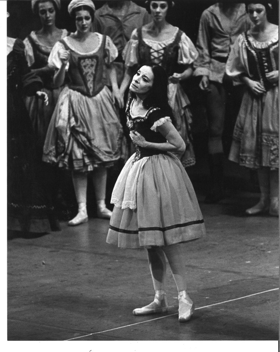 In this black and white photo, a dark haired ballerina in a peasant costume clutches her heart onstage.