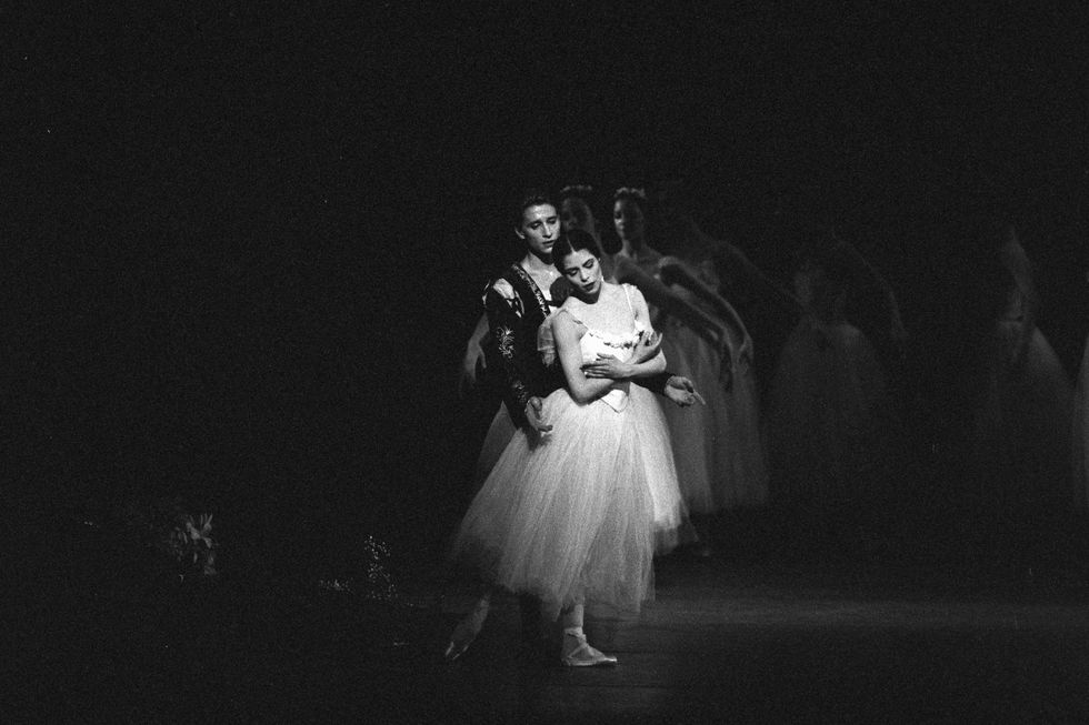 Susan Jaffe, wearing a white Romantic tutu, performs the role of Giselle, standing in tendu derriu00e9re on a darkened stage with her arms crossed low. Her partner, wearing a dark velvet tunic, stands right behind her, looking down at her.