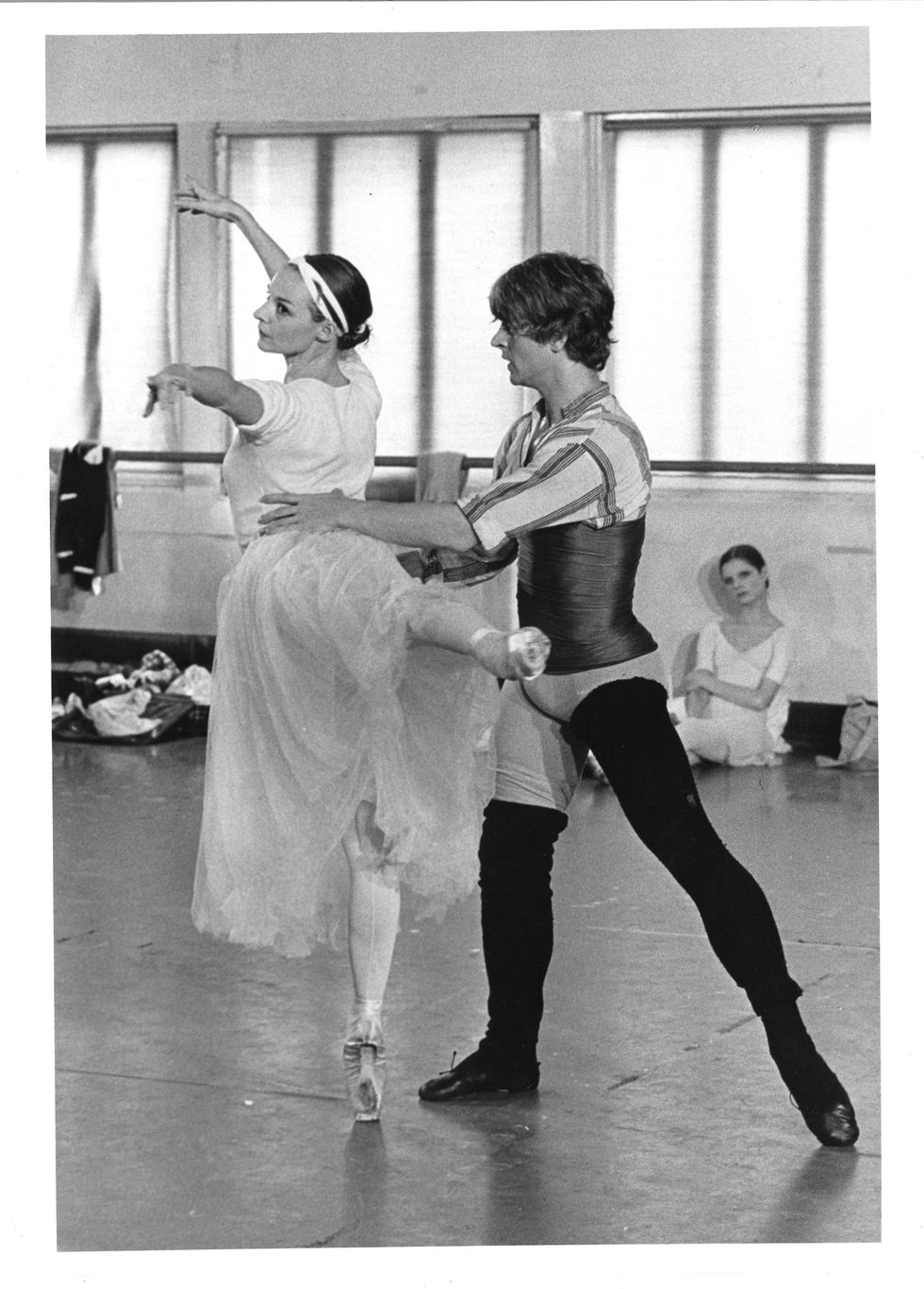 In a studio, Antoinette Sibley, wearing a t-shirt and long skirt over standard ballet practice clothes, piques to arabesque en pointe with her back to the camera, Mikhail Baryshnikov balancing her at the waist.