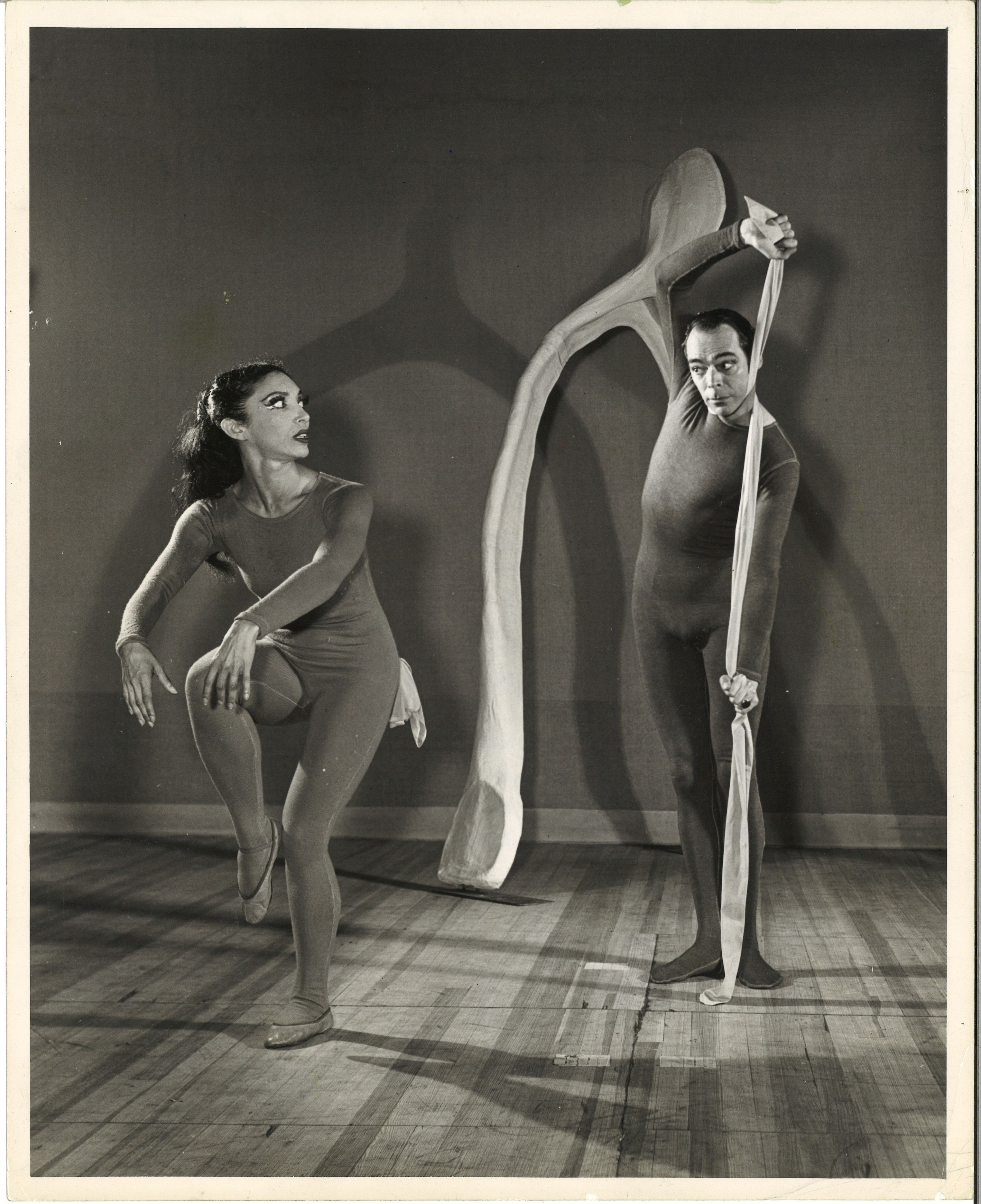 Two dancers are clothed in identical dark, tight-fitting, long-sleeved unitards. The woman balances on one leg, her arms curving over her raised knee, and looks over her shoulder with an expression of alarm at the man. He returns her look with raised eyebrows as he stretches a pale, thin length of fabric that is wrapped around his throat with both hands. In the background, a large set piece that looks like an oversized wishbone is propped against a wall.