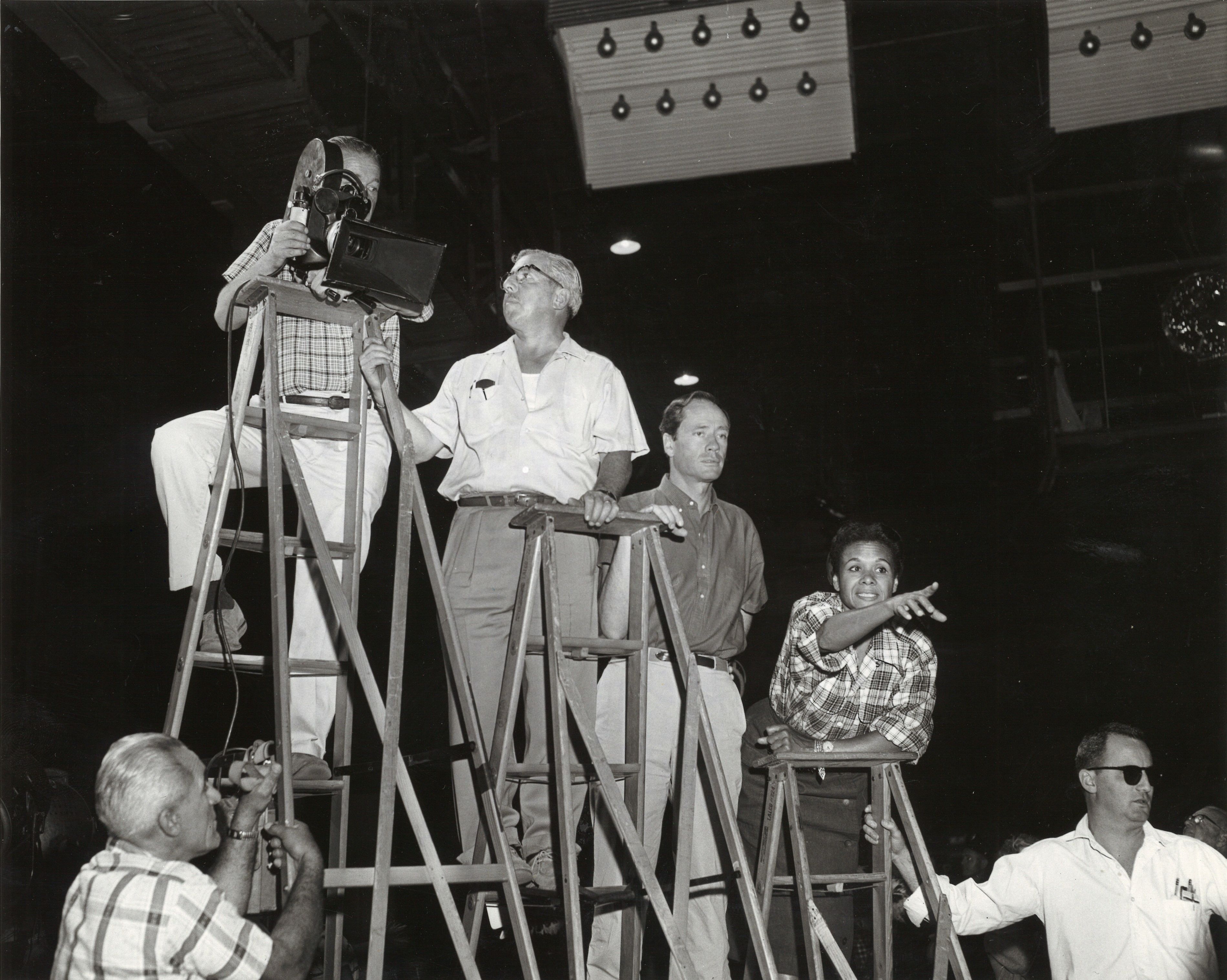 Dunham points as she stands atop a wooden latter. A white man follows her gaze, while a camera operator balances on a taller ladder, pointing the camera in the same direction.