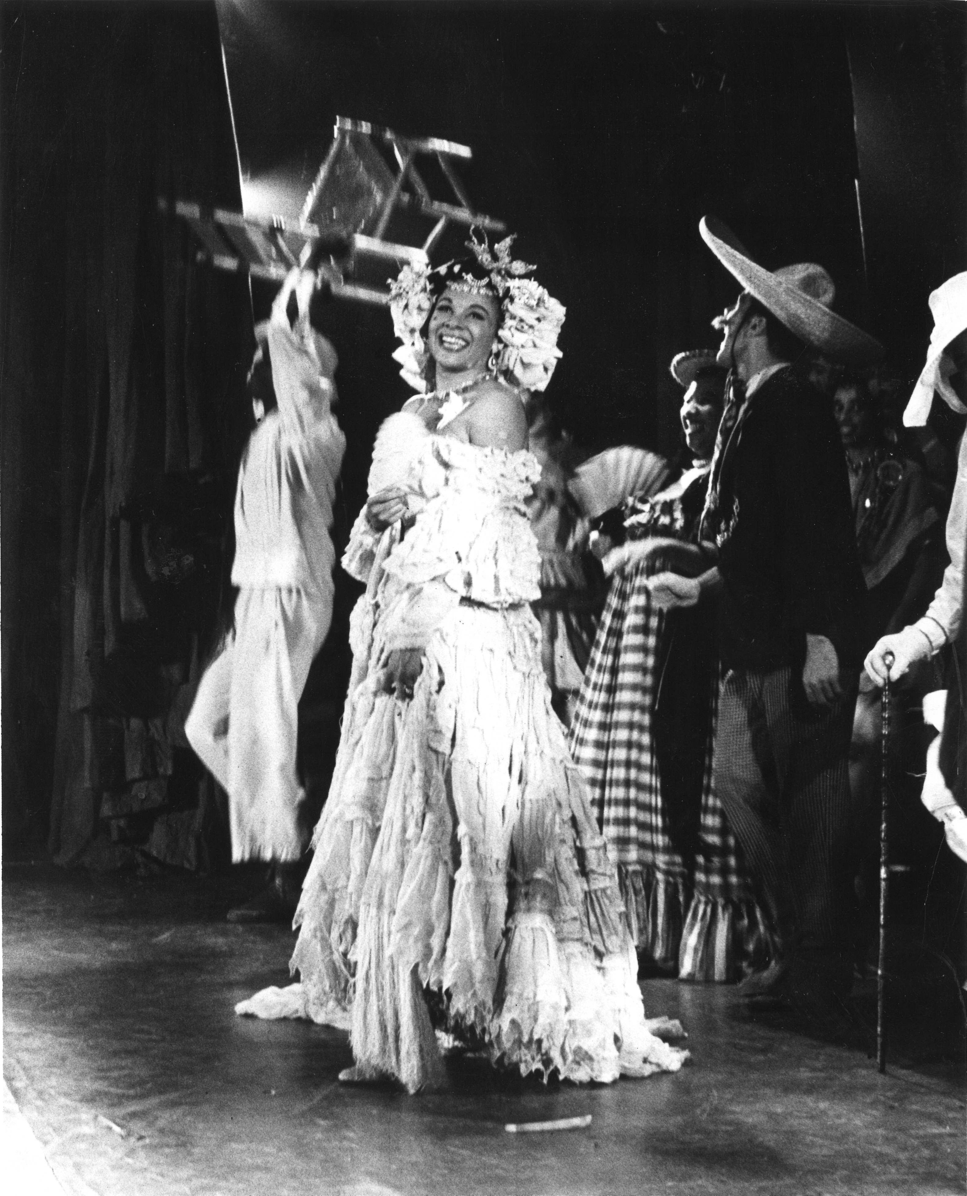 Katherina Dunham is captured onstage, mid-laugh, wearing an intricately draped off-the-shoulder dress and headdress. Other dancers are visible behind her, one of whom marches forward with a chair raised overhead.