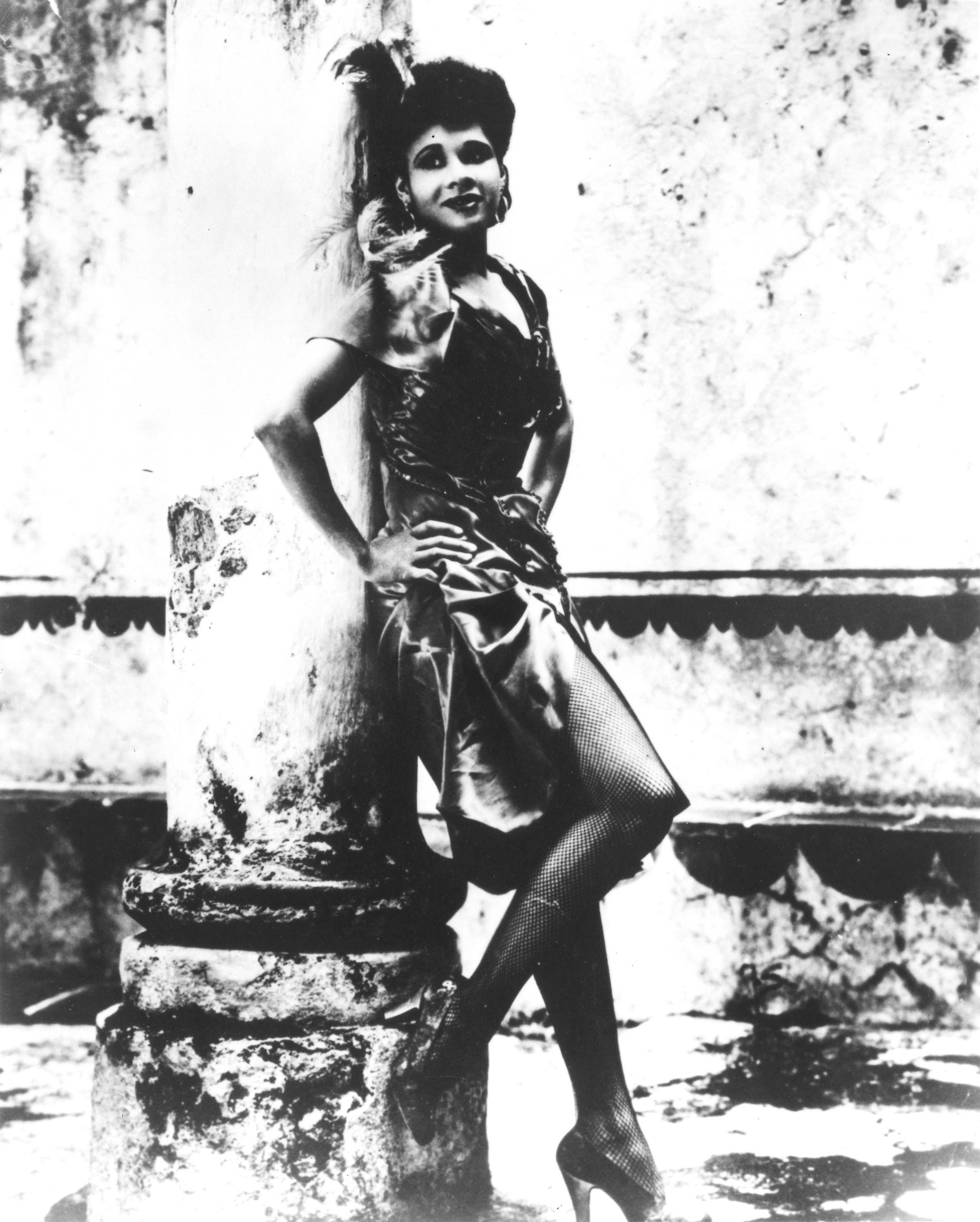A young Dunham smiles softly into the distance, posed against a pillar in a costume consisting of heels, fishnet tights, and a shiny, ruched dress.
