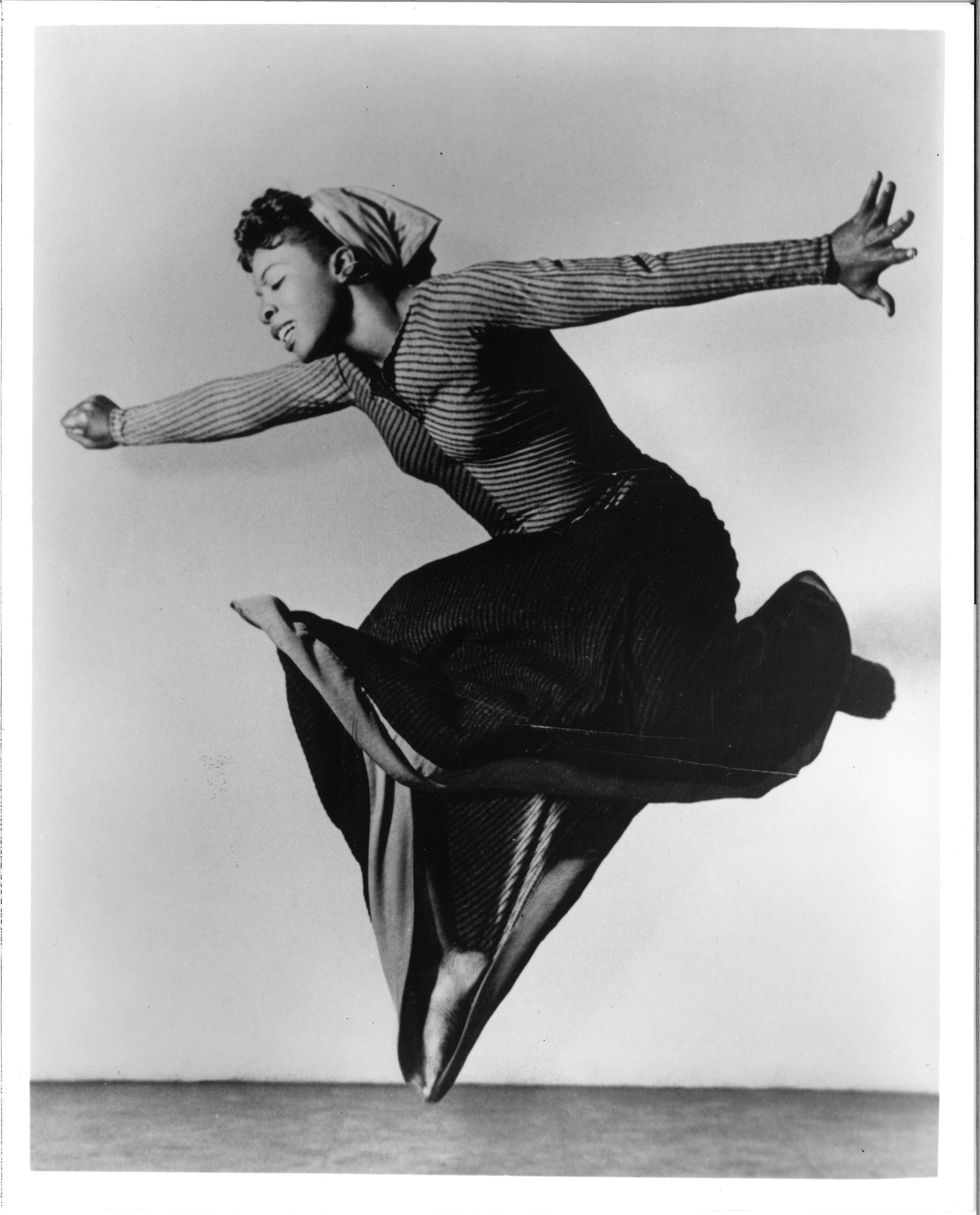 In an archival photo, Pearl Primus is caught midair. Her knees are tucked up underneath her, legs largely hidden by a black skirt. Her straight arms stretch before and behind her, the front hand in a fist, the back hand splayed wide. Her head is inclined toward the floor, eyes closed in an expression of joyous release.