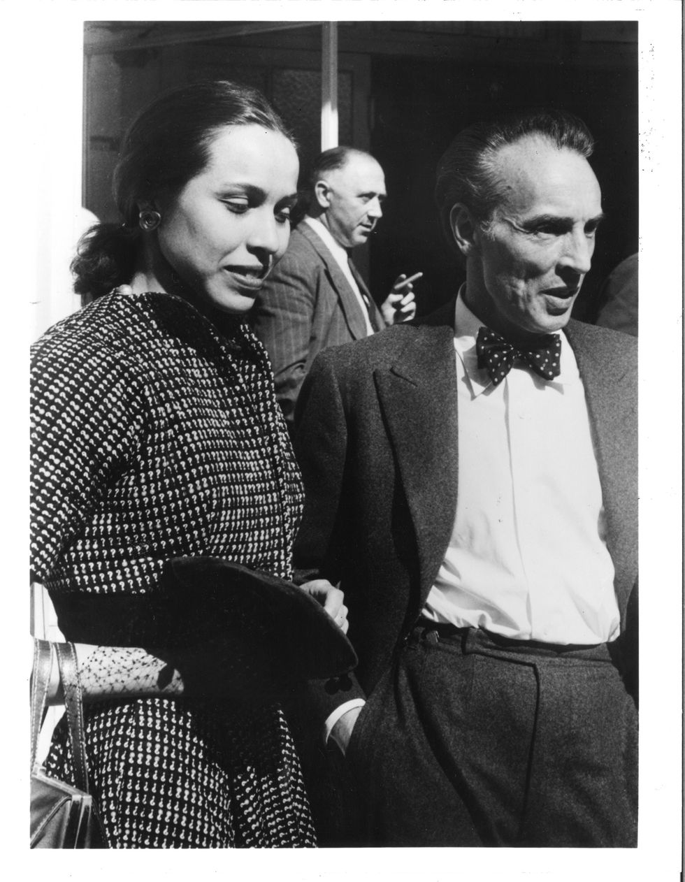 Maria Tallchief, wearing a nice patterned dress, and George Balanchine, in a suit with a bow tie, stand shoulder to shoulder, listening to someone speak off camera.