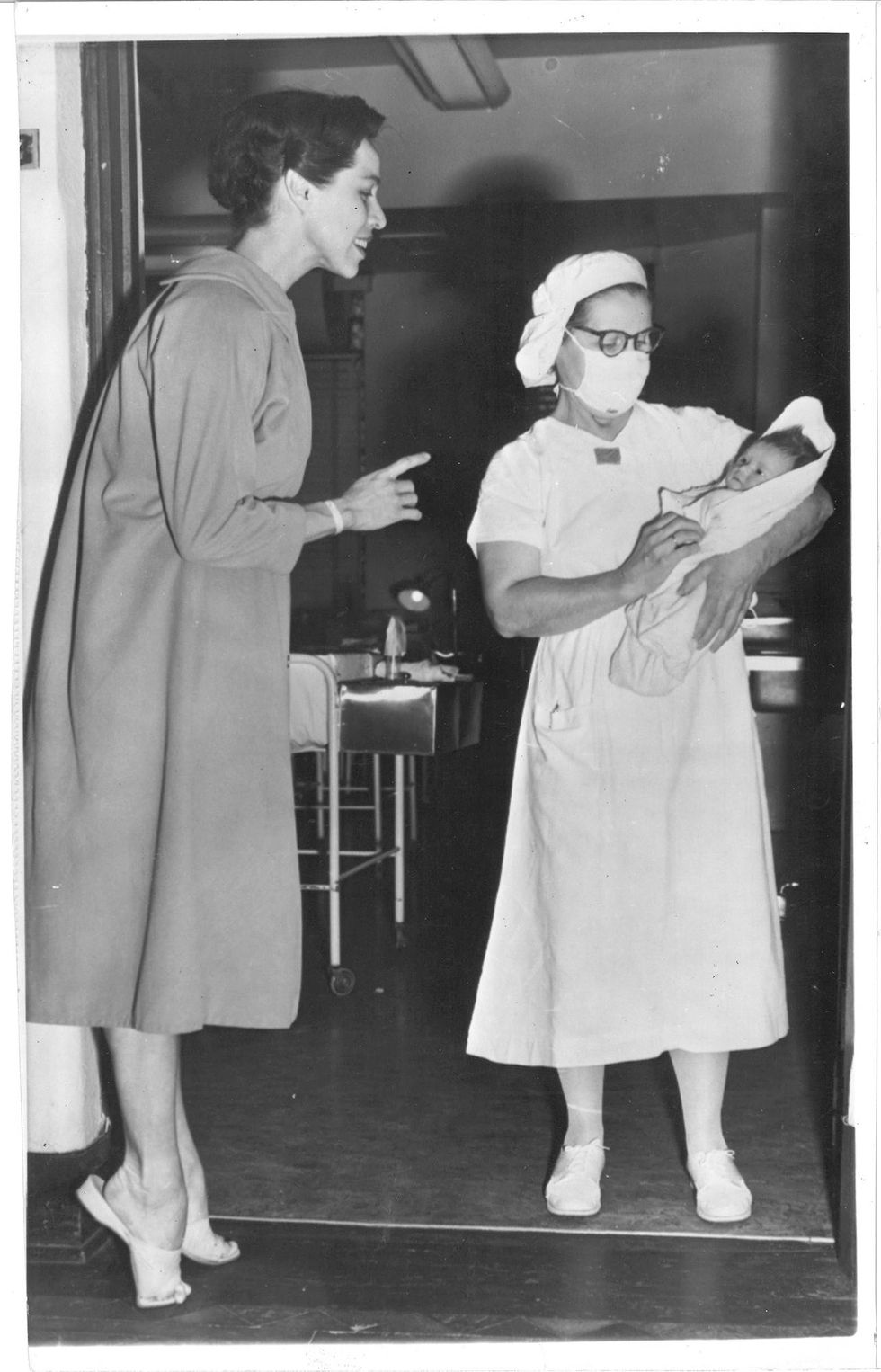Maria Tallchief relevu00e9s out of her heels and smiles in profile as she looks down at a newborn baby being held by a nurse in a white dress and face mask.