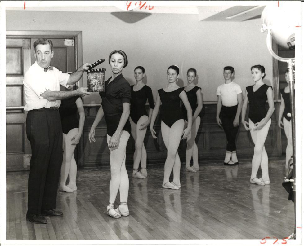 Maria Tallchief stands at the front of a wood-floored classroom in fifth position, arms in low fifth, as a man in shirtsleeves holds up a clapperboard next to her face. Behind her, younger dancers do the same.