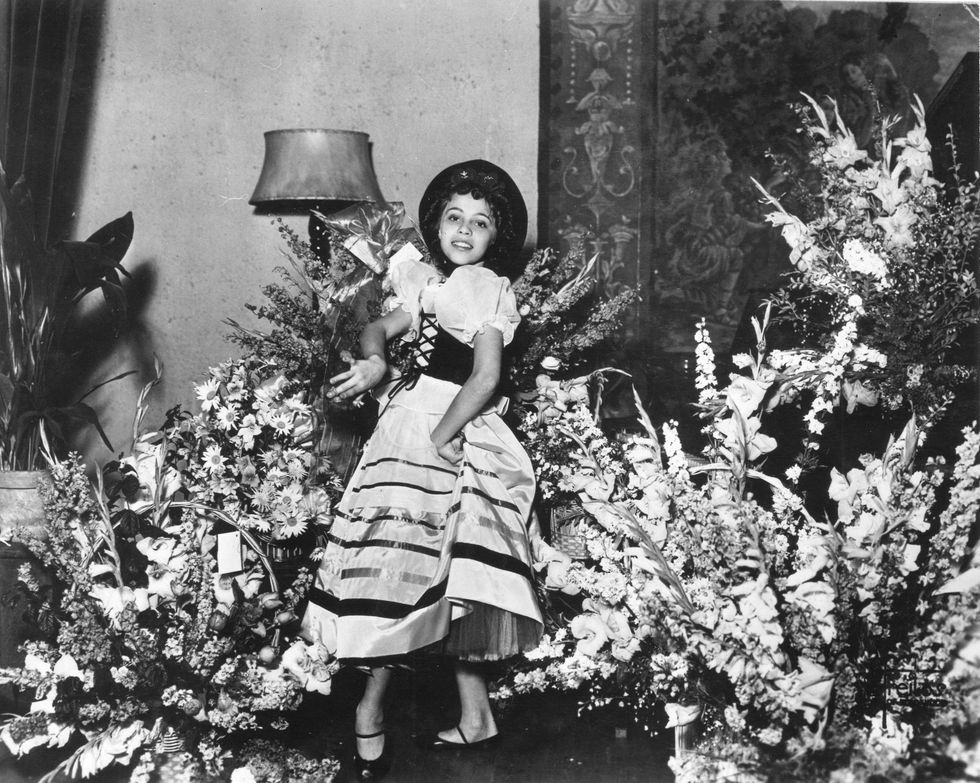 In a black and white image, a pre-teen Maria Tallchief poses in a room filled with flowers. She gathers the voluminous skirt of her poofy dress at one hip and curls her wrist as she holds castanets.