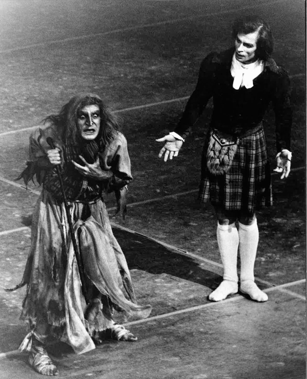 Erik Bruhn, in costume as Madge in a raggedy dress and wild, long wig wears a wild expression as he holds a clawed hand to his chest. A kilt-wearing Rudolf Nureyev watches curiously, palms open.