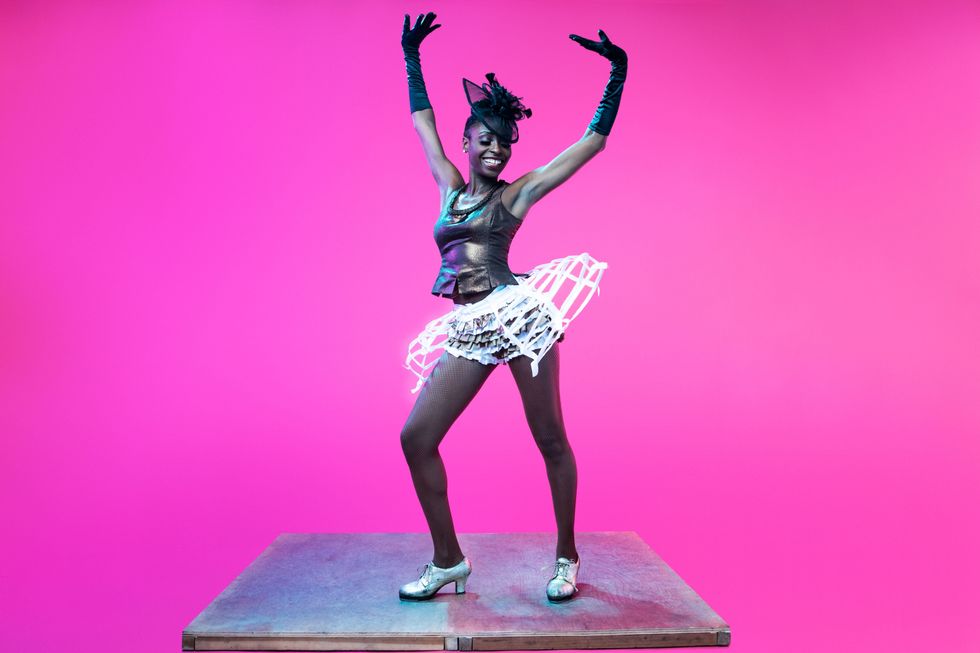 A dark skinned woman in silver, heeled tap shoes, a deconstructed, papery tutu, a shiny top, black gloves and a fancy black hat grins as she poses atop a silver surface floating on a bright pink background, arms flung into the air.