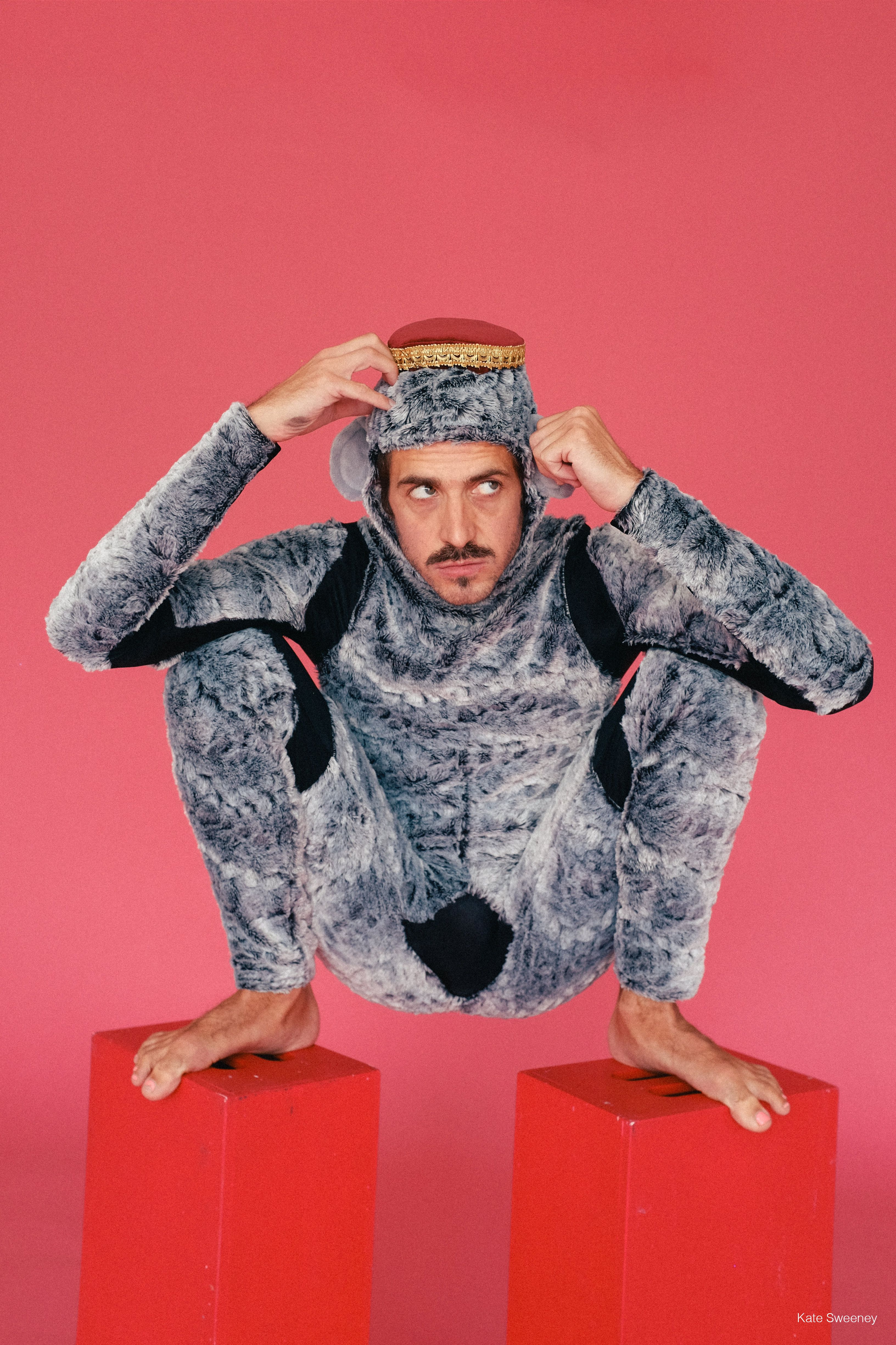 A man with a puzzled look dressed in a gray, fur-like jumpsuit, complete with monkey ears and a red fez, crouches atop two red blocks. His upper arms rest on his bent knees as his hands come to his temples.