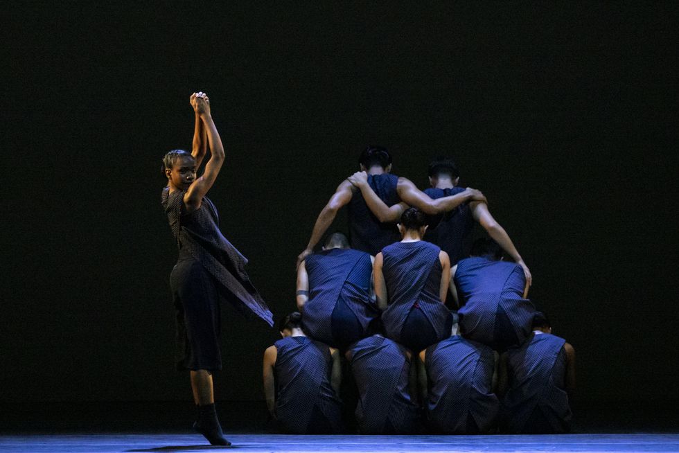 In the background, a pyramid of nine dancers sit facing backwards. In the foreground, a woman dancer stands on relevu00e9 with her hands clasped above her head.