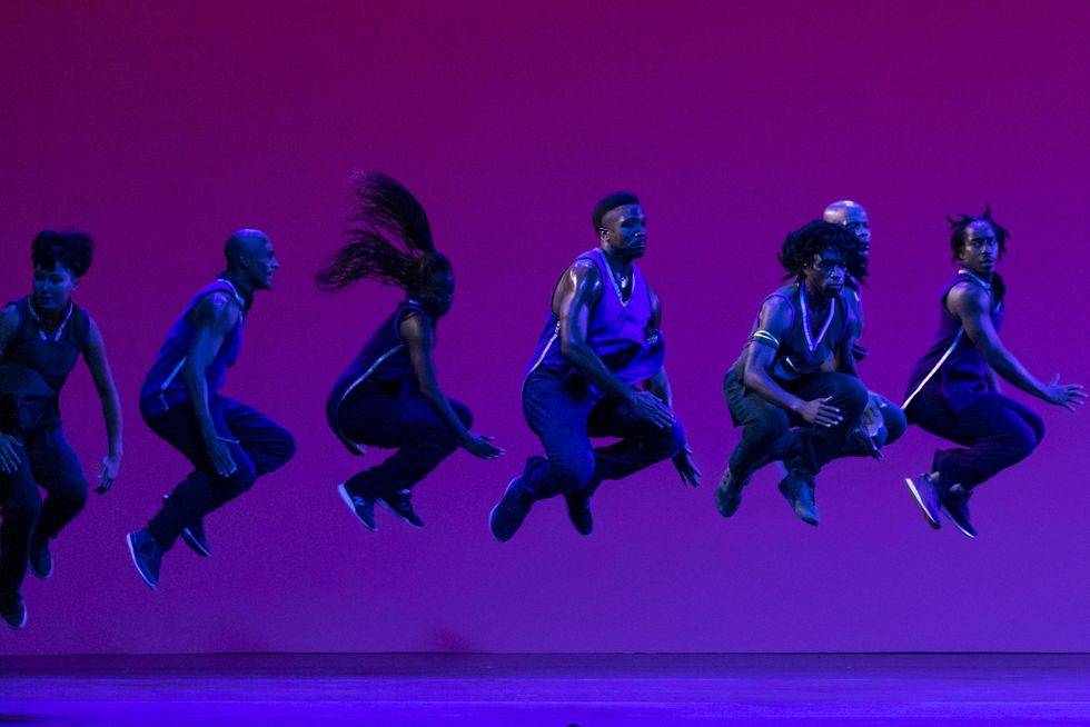 Seven dancers are captured jumping high in the air with their legs bent and tucked under their torsos.