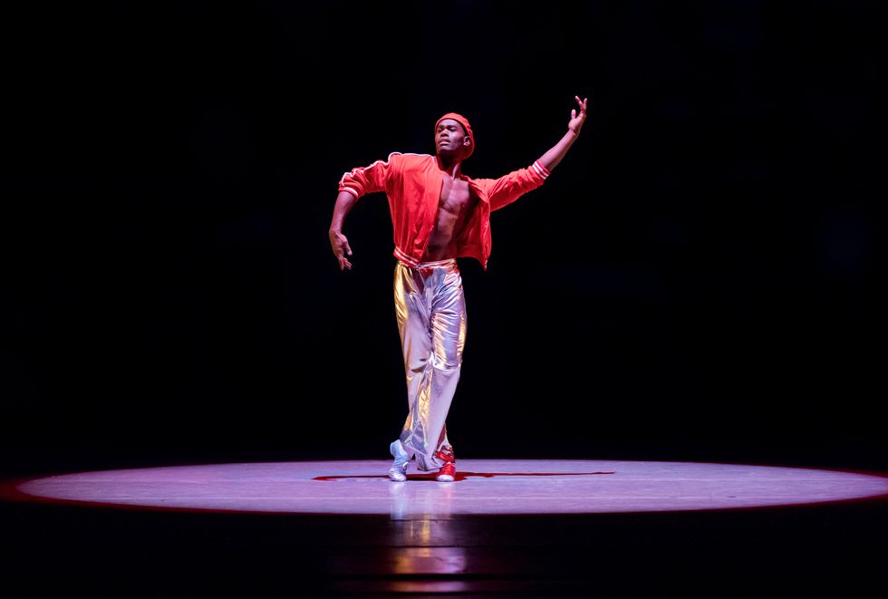 Roberts onstage in a spotlight. He wears shiny silver pants and an open red track jacket with no shirt underneath. He also wears a red cap. He crosses one leg in front of the other, and has his arms to the sides, one curved up and the other curved down. 