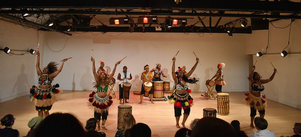 Four dancers stand in front of a group of drummers. They wear colorful traditional African attire. The dancers swing their arms up, holding drums sticks.
