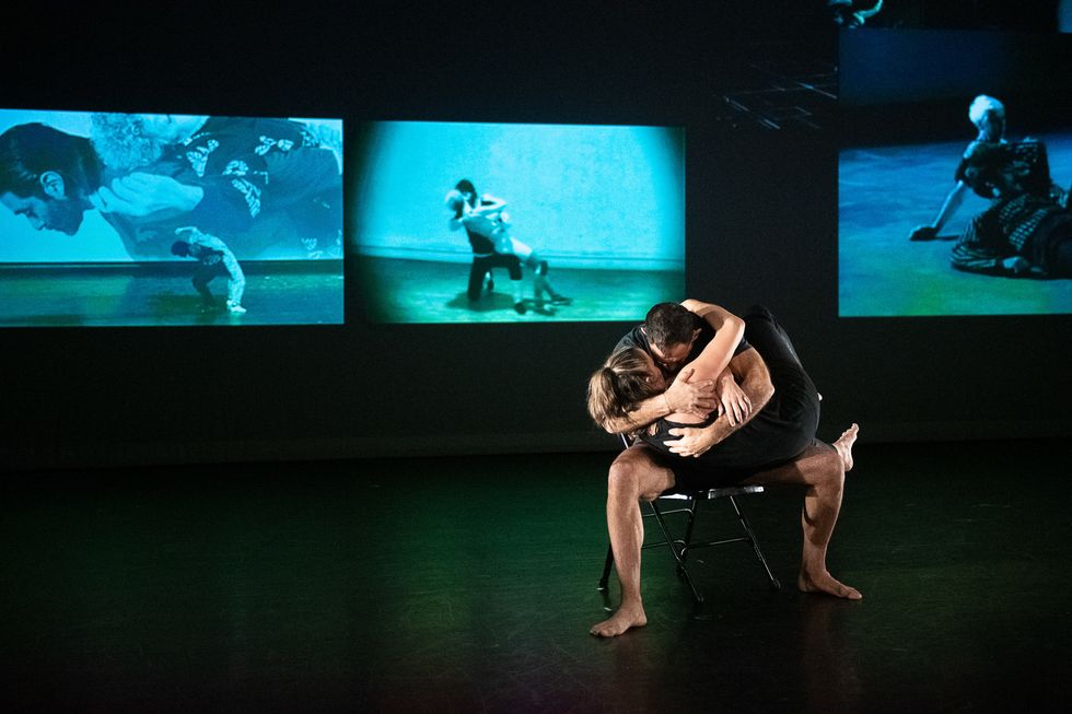 Two dancers on a chair embrace, in front of a project on the scrim.