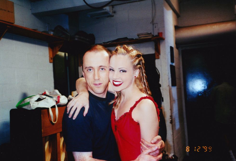 Amanda Schull poses behind the scenes with director Nicholas Hytner. The two hug, with Schull's arm around Hytner. Hytner is wearing a simple navy tee shirt, and Schull wears her costume, hair, and makeup from the finale of "Cooper's ballet"u2014red lipstick, cornrows, and a red dress.