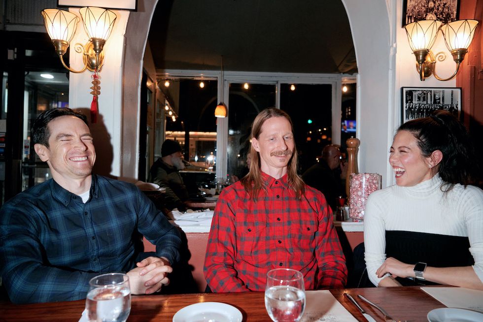 (From left) Radetsky, Stiefel, and Baiano seated at a dinner table. Radetsky and Stiefel both wear plaid, flannel button downs, and Baiano wears a black and white sweater. All three are caught mid-laughter.