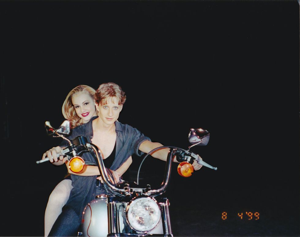 Schull and Stiefel pose on Stiefel's motorcycle. Stiefel wears an unbuttoned short sleeve shirt with a tank top beneath, and jeans. Both face the camera head-on, and smile.