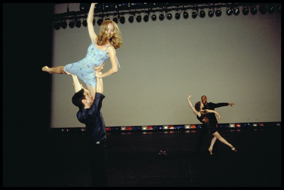 A still from "Center Stage." In the foreground, Radetsky lifts Schull above his head in variation of a press lift. Schull has one leg extended in arabesque, and the other pulled underneath her in passu00e9. She wears a light blue dress, with her hair down, and no shoes. Radetsky wears a simple black shirt and pants. In the background, we see two other dancers partnering.