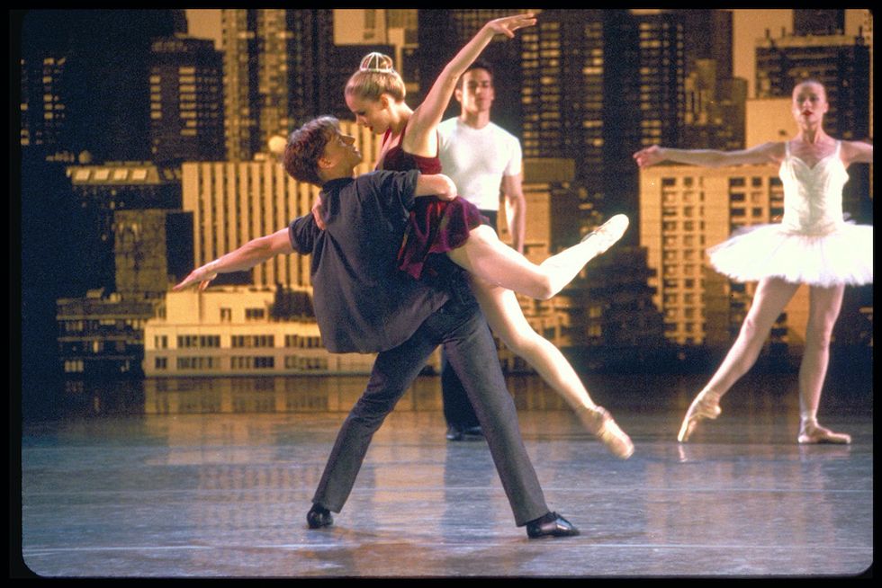 A still from "Center Stage." Stiefel lifts Schull. She wears a red dress, pink tights, pointe shoes, and her hair in a bun with a bedazzled bun cage. Stiefel wears black jeans, a black tee shirt, and black jazz shoes. They look into each other's eyes.