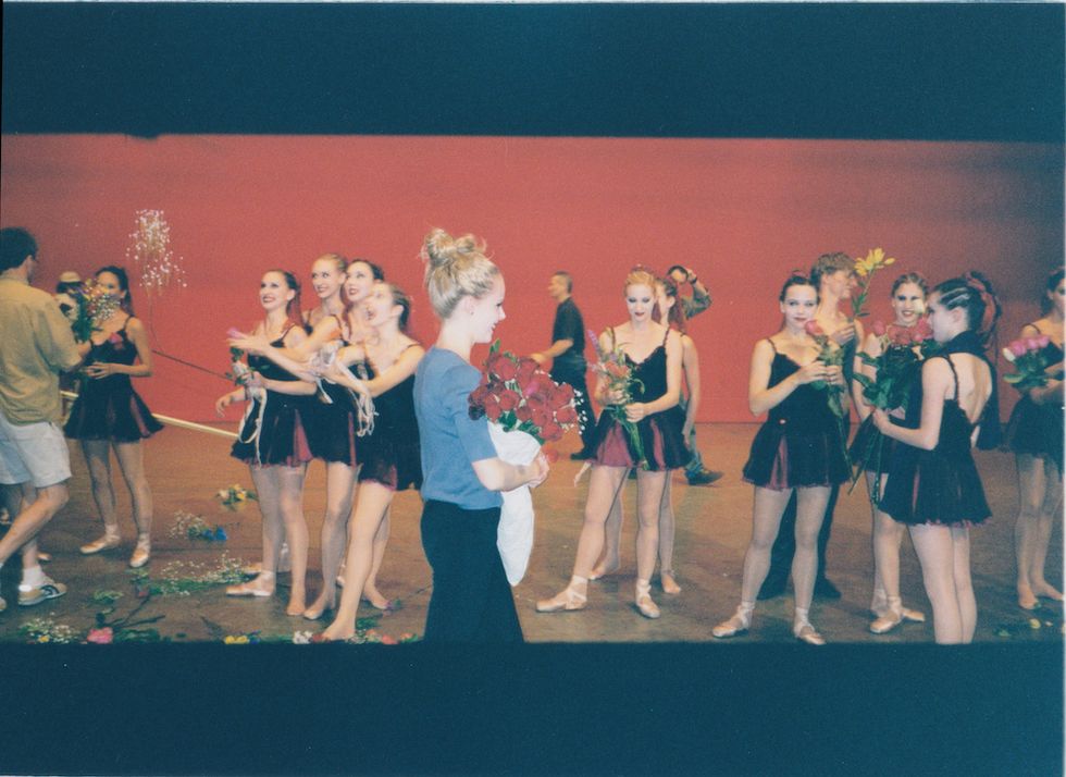 A candid photo of Schull behind the scenes. She holds a bouquet of red roses, and wears a blue sweater with black pants. Other dancers from the film stand casually in the background, wearing their costumes from "Cooper's ballet," also holding flowers.