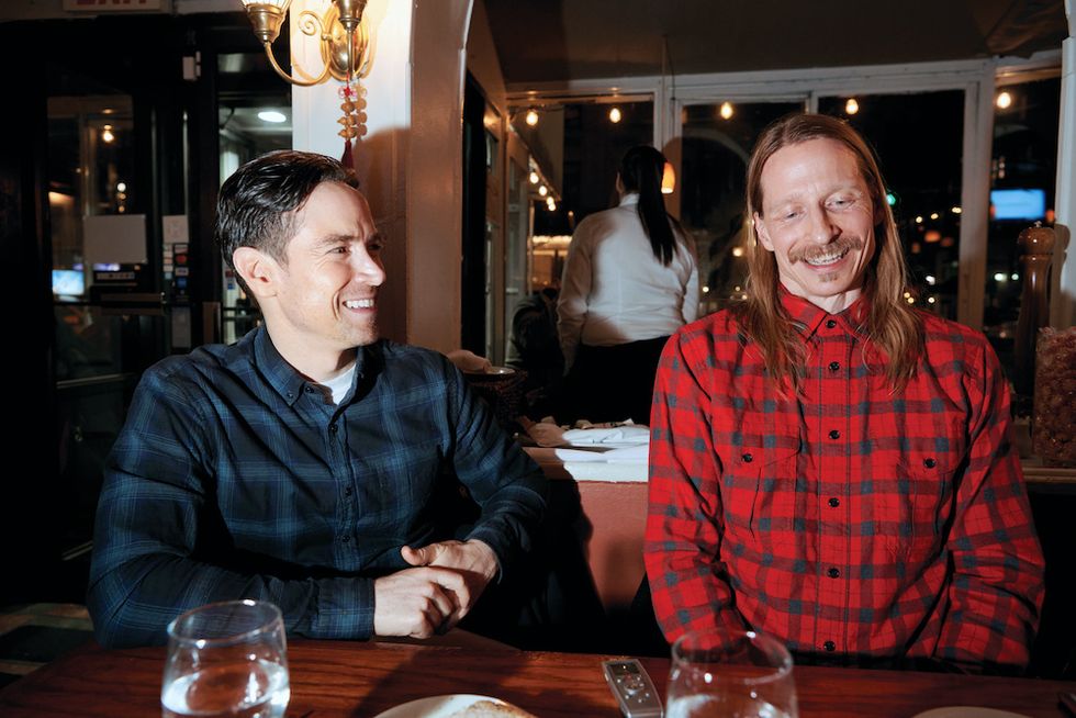 Sascha Radetsky and Ethan Stiefel pose side by side, seated at a restaurant. Both are wearing flannel button-downs, and both are caught mid-laughter.