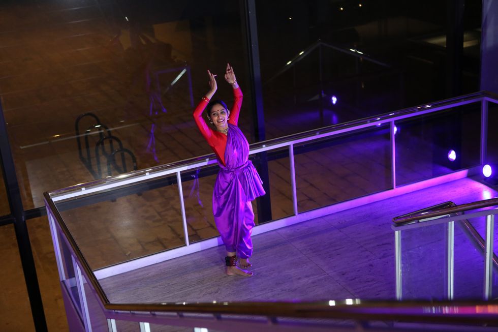 A smiling dancer in purple and red poses with her arms above her head, hands shaping a specific mudra, on a landing between staircases. She is surrounded by glass.