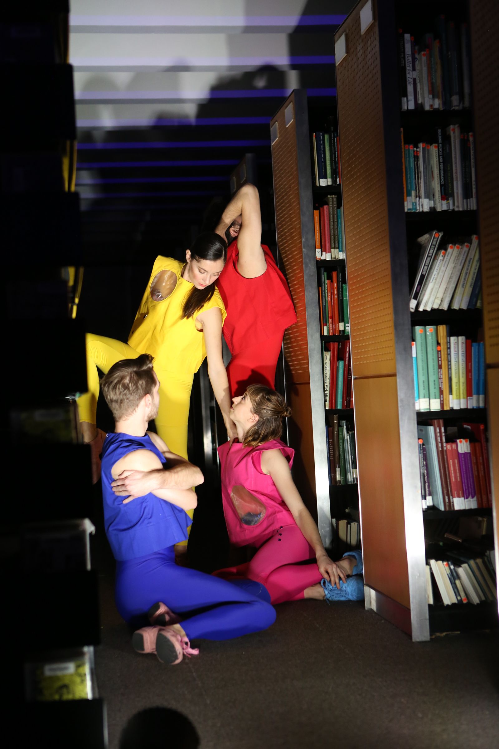 Four dancers in red, yellow, blue, and pink pose in a narrow aisle between bookshelves. The two closest to the front are seated on one hip, looking up at the other two. The one in yellow balances on one leg, her hand resting atop one of her fellow's shoulders. Behind, a tall dancer in red stands facing a shelf, arm lifted to hide his face from view.