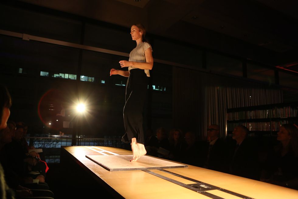 A woman atop a long table is lit from the side in an otherwise dark room. She is barefoot, rising onto the ball of one foot as the other arcs up from striking the floor. Her focus is ahead and slightly down, wholly focused on what she is doing.